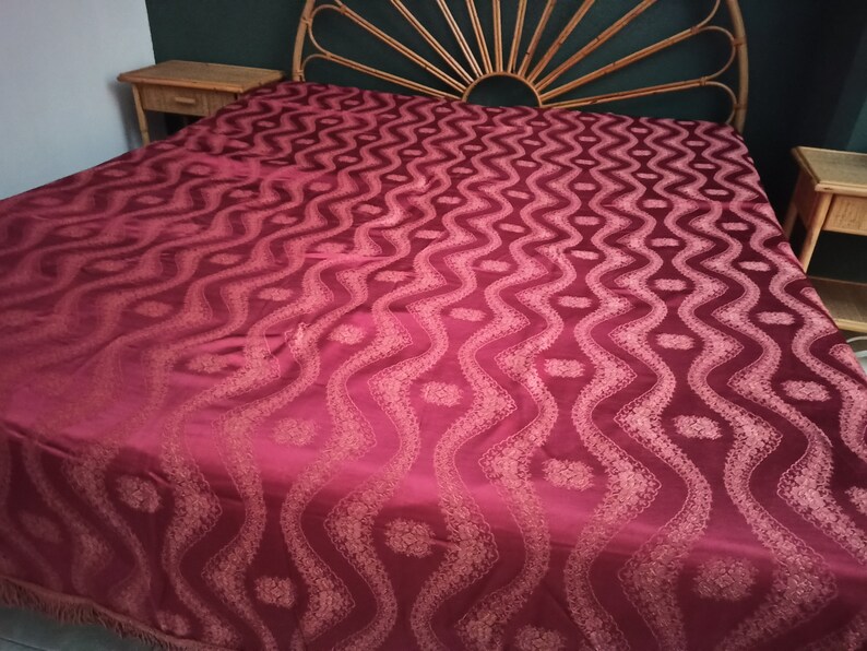 May include: A burgundy bedspread with a repeating wave pattern and floral accents. The bedspread is draped over a bed with a woven headboard and two matching nightstands. The fabric has a subtle sheen, suggesting a luxurious material.