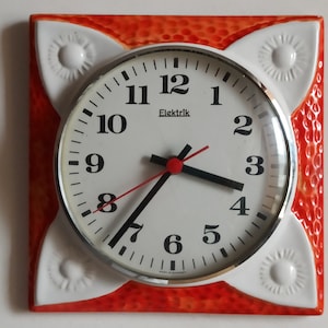 May include: A vintage wall clock with a white face and black numerals, the clock hands are black and red. The clock is set in a square frame with a textured orange background and white decorative accents. The word "Elektrik" is printed on the clock face.