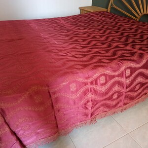 May include: A burgundy bedspread with an intricate wave pattern and a fringed edge. The bed covering is draped over a bed frame, showcasing its detailed design and texture. The color is a deep, rich shade.