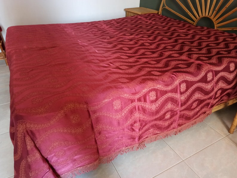 May include: A burgundy bedspread with an intricate wave pattern and a fringed edge. The bed covering is draped over a bed frame, showcasing its detailed design and texture. The color is a deep, rich shade.