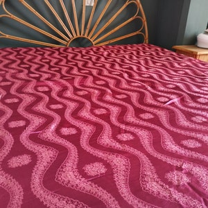 May include: A bed with a burgundy patterned bedspread. The bed has a bamboo headboard and two matching nightstands. An iron sits on one of the nightstands. The bedspread features a repeating wave-like design with floral accents.