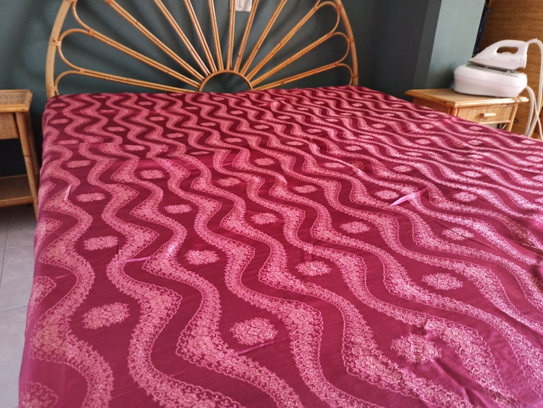 May include: A bed with a burgundy patterned bedspread. The bed has a bamboo headboard and two matching nightstands. An iron sits on one of the nightstands. The bedspread features a repeating wave-like design with floral accents.