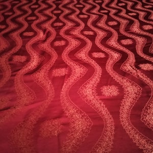 May include: A deep red fabric with a repeating pattern of wavy lines and floral accents. The design is embroidered with a lighter shade of red, creating a textured and elegant appearance. The fabric has a soft sheen.