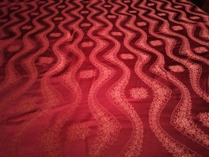 May include: A deep red fabric with a repeating pattern of wavy lines and floral accents. The design is embroidered with a lighter shade of red, creating a textured and elegant appearance. The fabric has a soft sheen.