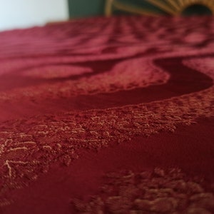 May include: Close-up of a deep red fabric with a swirling, textured pattern. The fabric has a subtle sheen and a detailed, raised design in a lighter shade of red, creating a sense of depth and luxury. The overall theme is textiles.