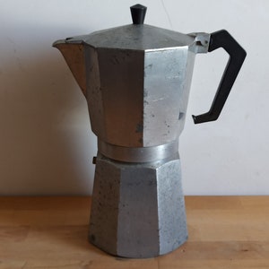Op de afbeelding: Een zilverkleurige espressomaker voor op het fornuis met een zwarte handgreep en knop. De koffiepot is gemaakt van metaal en heeft een klassiek, achthoekig ontwerp. Hij staat op een houten oppervlak tegen een witte muur.