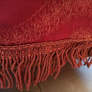 May include: Close-up of a burgundy fabric with a detailed, golden floral pattern. The fabric is trimmed with a matching twisted rope and fringe. The texture and color suggest a decorative textile, possibly for upholstery or home decor.
