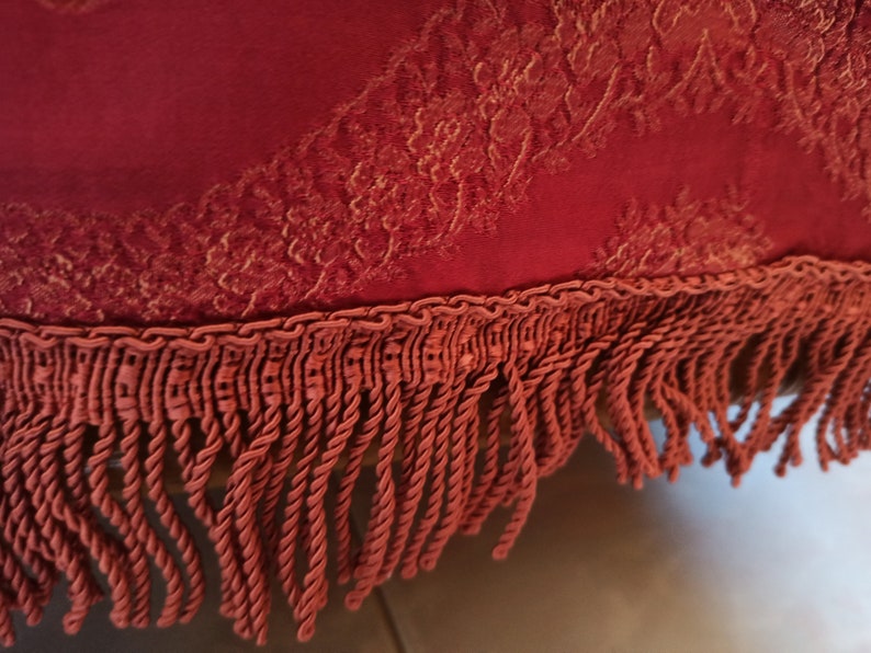 May include: Close-up of a burgundy fabric with a detailed, golden floral pattern. The fabric is trimmed with a matching twisted rope and fringe. The texture and color suggest a decorative textile, possibly for upholstery or home decor.