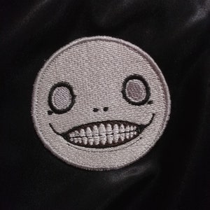 May include: A white embroidered patch with a smiling face with two large eyes and a toothy grin. The patch is on a black background.