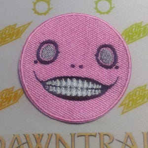 May include: Pink embroidered patch with a smiling face with large eyes and a toothy grin.