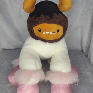 May include: A plush toy with a white body, brown fur, and orange horns. The toy has a black face with a toothy grin and pink sparkly shoes.