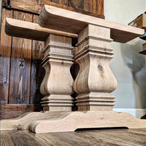 May include: A light-colored, unfinished wooden table base with two ornate pedestal supports. The base features a wide, curved foot and a thick tabletop support. The wood grain is visible, and the design is rustic and elegant.