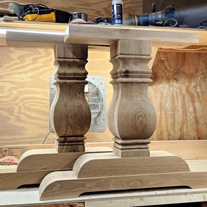 May include: Two unfinished wooden table bases. Each base has a rectangular top, a decorative, turned pedestal, and a rectangular foot. The wood grain is visible. The bases are in a workshop setting.