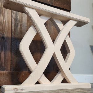 May include: A light-colored wooden console table with a geometric base. The table features a rectangular top supported by an intricate, interwoven diamond-shaped design. The wood has a natural finish, showcasing the grain.