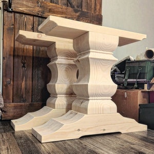 May include: Two unfinished wooden table legs with a pedestal base and a curved bottom. The legs are made of light-colored wood and have a natural finish.