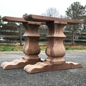 May include: Two wooden table bases with a unique, curved design. The bases are made of dark wood with a natural grain pattern. The bases are connected to each other and have a square top.