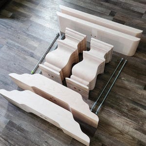 May include: Unassembled wooden table legs with metal rods and screws. The legs are made of light wood and have a curved design. The metal rods are silver and are used to connect the legs to the table top.