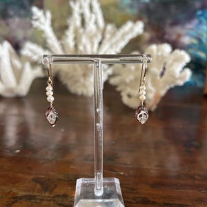 May include: A pair of dangle earrings displayed on a clear stand. Each earring features a gold-colored hook, a strand of small white pearls, and a teardrop-shaped charm with a dark purple and clear design. The background includes white coral and a blurred blue and green backdrop.