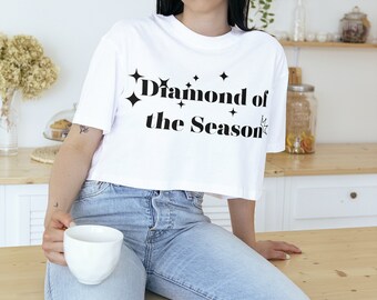 May include: A white cropped t-shirt with the text "Diamond of the Season" in black lettering and small star accents. The shirt is worn by a person sitting on a wooden counter.