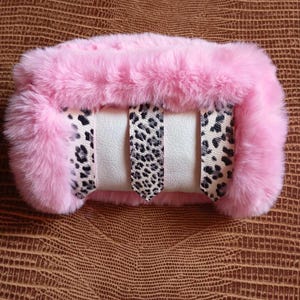 Puede incluir: Un brazalete de piel sintética rosa con una sección central de color crema y detalles de estampado de leopardo. El brazalete está diseñado para usarse alrededor de la muñeca y está hecho de materiales suaves.