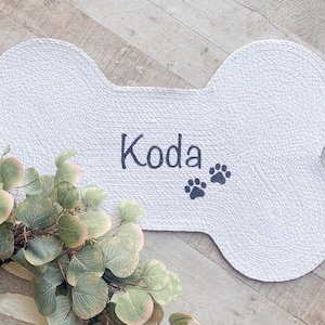 Puede incluir: Alfombra blanca para mascotas con forma de hueso con el nombre "Koda" y dos huellas de patas bordadas en negro.