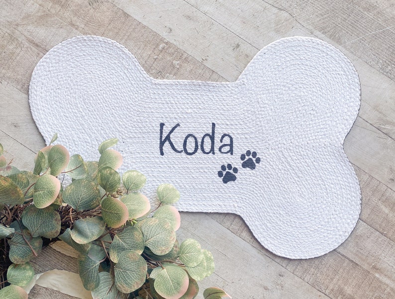 Puede incluir: Alfombra blanca para mascotas con forma de hueso con el nombre "Koda" y dos huellas de patas bordadas en negro.