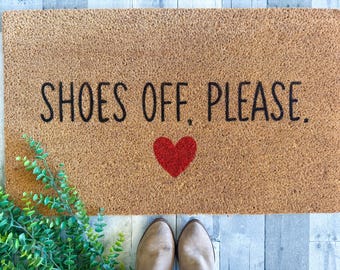 May include: A rectangular doormat made of natural fibers, featuring the text "SHOES OFF, PLEASE." in black, with a red heart. Green leafy plant on the left side. Light brown shoes are visible at the bottom.