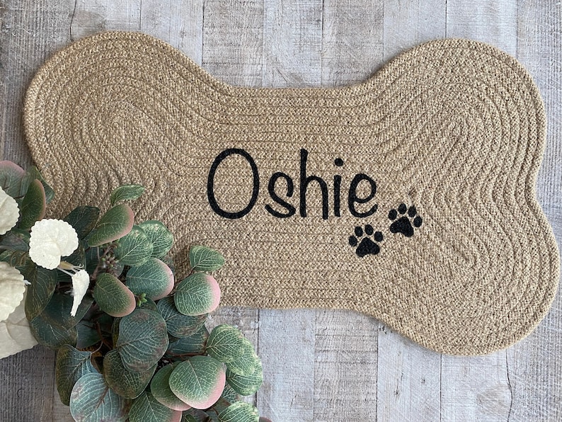 Puede incluir: Una alfombra de bienvenida de color beige en forma de hueso hecha de cuerda trenzada con el nombre "Oshie" y dos huellas de patas negras bordadas.