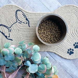 Puede incluir: Una alfombra para perros de color beige en forma de hueso con una silueta de perro negra y huellas de patas. Un cuenco de madera lleno de comida para perros marr&oacute;n se encuentra sobre la alfombra.