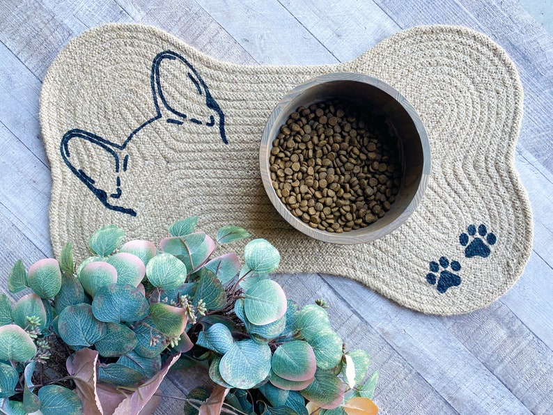 Puede incluir: Una alfombra para perros de color beige en forma de hueso con una silueta de perro negra y huellas de patas. Un cuenco de madera lleno de comida para perros marr&oacute;n se encuentra sobre la alfombra.
