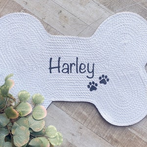 Puede incluir: Cama para perro de hueso blanco con el nombre "Harley" bordado en negro y dos huellas de patas negras.