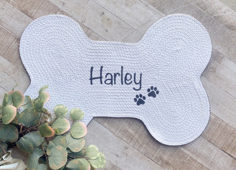 Puede incluir: Cama para perro de hueso blanco con el nombre "Harley" bordado en negro y dos huellas de patas negras.