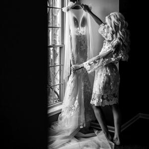 May include: A black and white image of a wedding dress hanging by a window. The dress is white with floral lace detailing and a long train. A sheer veil is draped over the dress. A person is holding the dress, wearing a matching lace robe.