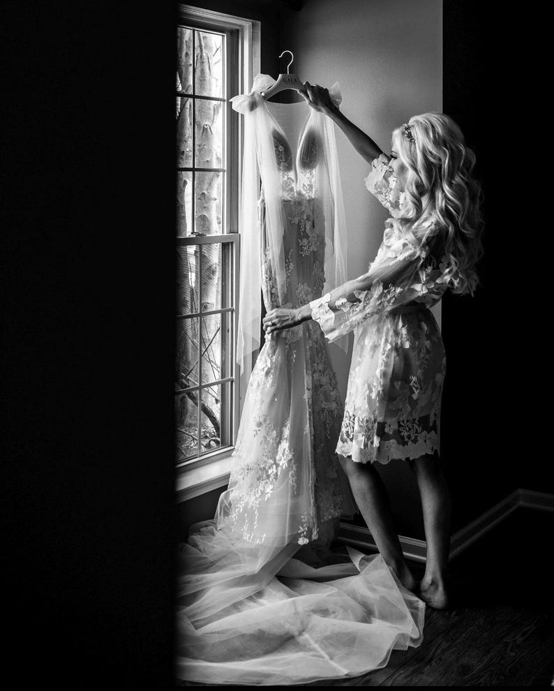 May include: A black and white image of a wedding dress hanging by a window. The dress is white with floral lace detailing and a long train. A sheer veil is draped over the dress. A person is holding the dress, wearing a matching lace robe.