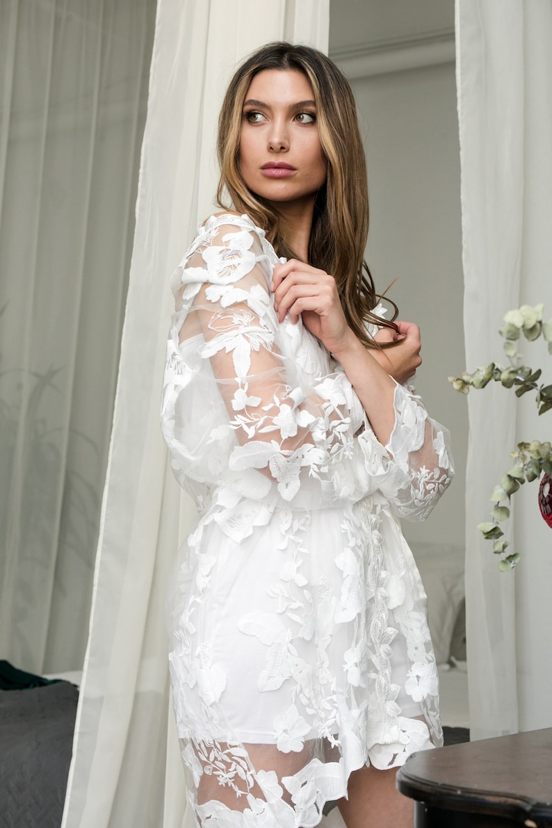 May include: A white, knee-length dress with floral lace overlay. The dress features long, sheer sleeves with floral detailing and a matching floral pattern on the bodice and skirt. The model is standing near a sheer white curtain.