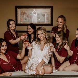 May include: A group of women in matching burgundy pajamas with white trim, toasting with drinks. The bride is in a white lace robe. The setting is a bedroom with a framed picture on the wall.