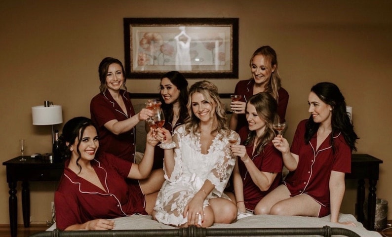 May include: A group of women in matching burgundy pajamas with white trim, toasting with drinks. The bride is in a white lace robe. The setting is a bedroom with a framed picture on the wall.