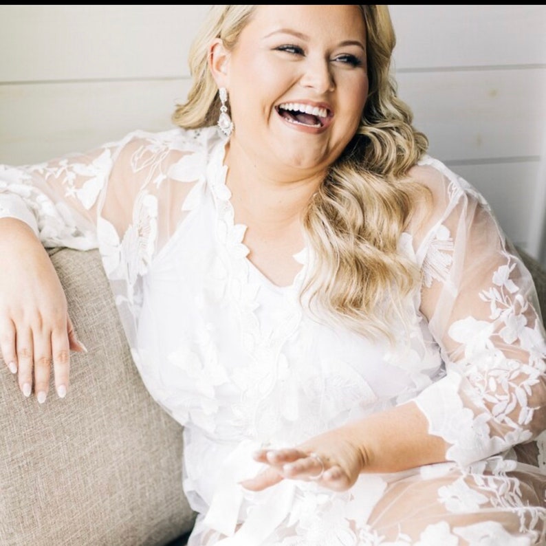 May include: A woman wearing a white, floral lace robe, smiling and laughing. The robe has a V-neck and long sleeves with floral detailing. She is sitting on a gray couch. Her blonde hair is styled in loose waves, and she wears dangling earrings.