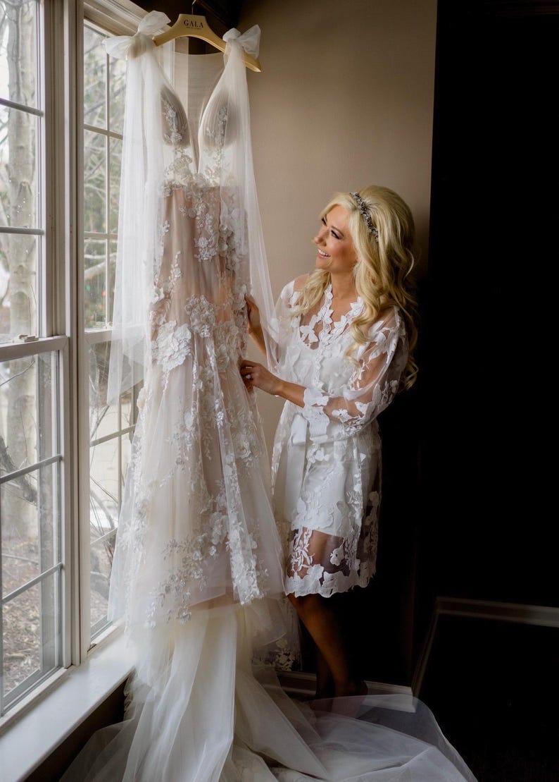 May include: A white wedding dress with floral lace detailing hangs near a window. The dress is on a wooden hanger with the word "GALA" printed on it. A woman in a white lace robe stands nearby.