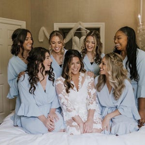 May include: A group of seven women, smiling and laughing, are gathered together. Most are wearing light blue robes with ruffled sleeves. One woman is wearing a white, lacy robe. A wedding dress hangs in the background, suggesting a bridal theme.