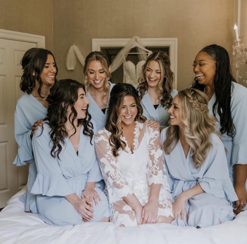 May include: A group of seven women, smiling and laughing, are gathered together. Most are wearing light blue robes with ruffled sleeves. One woman is wearing a white, lacy robe. A wedding dress hangs in the background, suggesting a bridal theme.