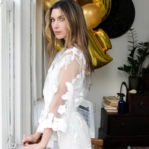May include: A white, sheer robe with floral embroidery. The robe features a belted waist and wide sleeves. The model is standing near a window with gold balloons in the background. The robe is a delicate and elegant piece.