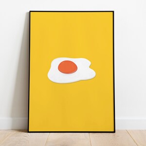 May include: A framed art print with a minimalist fried egg illustration. The egg has a white, irregular egg white and a round, orange yolk, set against a solid yellow background. The artwork is presented in a black frame.