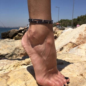 May include: A black beaded anklet with silver beads. The anklet is layered and is worn on a person's ankle.