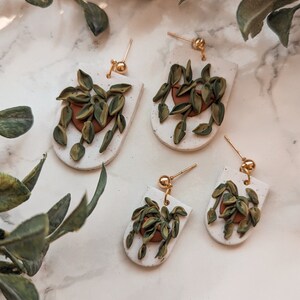 May include: Four pairs of white speckled clay earrings with a gold post and ball. Each earring features a brown pot with green plant leaves. The earrings are in a half-moon shape.