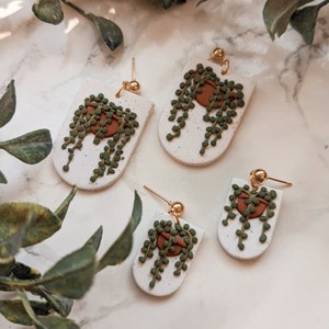 May include: Handmade polymer clay earrings featuring a string of pearls plant design. The earrings have a white speckled background with a brown pot and green plant details. The earrings have gold-colored ear wires.