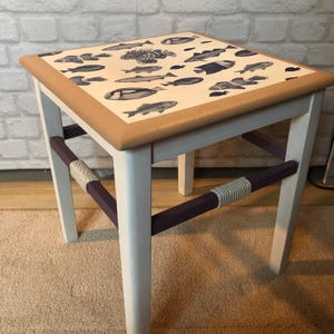 Könnte beinhalten: Ein quadratischer Beistelltisch mit einer hellbraunen Tischplatte, die mit einem dunkelblauen Fischmuster verziert ist. Der Tisch hat vier weiße Beine, die durch violette Stützstreben verbunden sind, welche mit hellem Seil umwickelt sind.