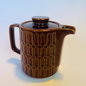 Op de afbeelding: Een bruine keramische theepot met een deksel en een handvat. De theepot heeft een geometrisch patroon en een tuit om uit te schenken. Geschikt voor het serveren van thee of andere warme dranken.