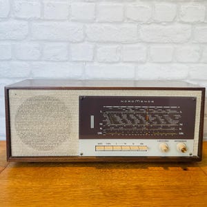 May include: A vintage Nordmende radio featuring a rectangular wooden cabinet, a textured cream-colored speaker grille, and a dark brown glass display panel with frequency scales. It includes five rectangular push buttons and two circular knobs.