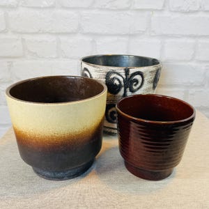 Può includere: Tre vasi di ceramica di varie dimensioni e colori. Il vaso più grande ha una sfumatura da beige a marrone. Il vaso centrale è bianco con disegni a spirale neri. Il vaso più piccolo è di un marrone rossiccio scuro.